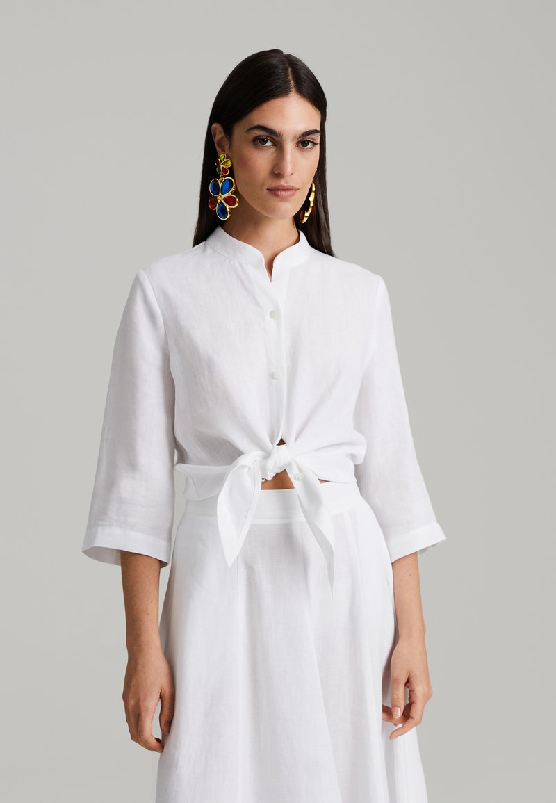 Bela lanena srajca z mandarin ovratnikom, zavezana v pasu. Dolgi rokavi z zavihanimi manšeti. Model nosi barvite, velike cvetlične uhane.