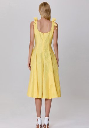 Robe jaune avec une finition texturée, sans manches, et des bretelles à nouer. Présente un dos dégagé et une silhouette en A, longueur genou.