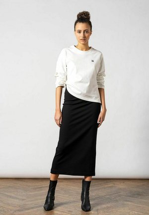 Weißes Langarmhemd mit kleinem Logo, kombiniert mit einem taillierten schwarzen Midirock und schwarzen Stiefeletten; glatte Texturen, schlichtes Design.