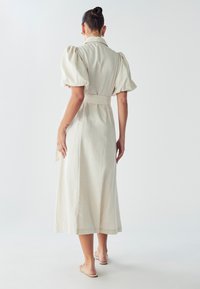 Šviesiai smėlio spalvos midi suknelė su pūstomis rankovėmis ir surišta juosmeniu, turinti apykaklę ir plasdantį sijoną. Paprastas, tekstūruotas audinys.