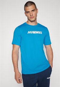 Camiseta de algodón de manga corta en color azul, con un logo blanco de "HUMMEL" en el pecho. Textura suave, cuello redondo.