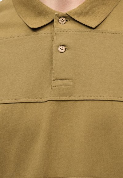 Πράσινο πουκάμισο πόλο με γιακά, δύο κουμπιά κλειστά στην κορυφή και ορατή οριζόντια ραφή κατά μήκος του στήθους.