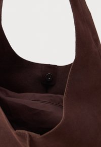 Bolsa tote em camurça castanha com um formato curvado e uma abertura ampla, apresentando um fecho de pressão e um bolso interior com fecho éclair. Textura suave em toda a parte.