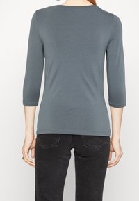 Grå topp med 3/4 ärm i ett slätt, elastiskt tyg. Har rund halsringning och en smal passform, ihop med mörka jeans.