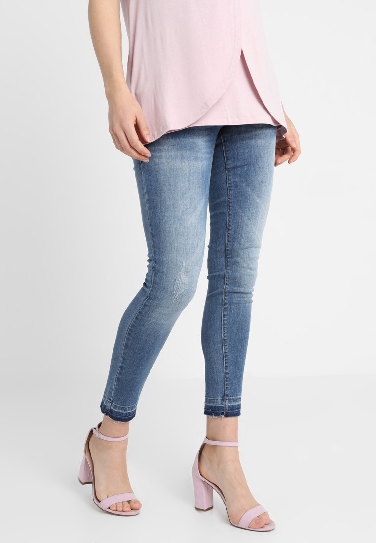 Kvinna som bär blekta blå skinny jeans, en ljusrosa topp och ljusrosa klacksandaler med blockklack, stående mot en enkel vit bakgrund.