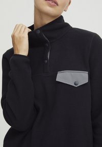 Sort fleece pullover med høj krave, trykknapper og en grå brystlomme. Blød tekstur med funktionelle design detaljer.