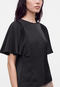 Femme portant un haut noir avec de larges manches flottantes et un col rond, présentée des épaules à la taille sur un fond uni.