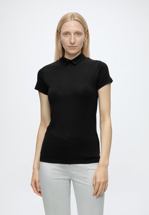 Femme aux cheveux blonds raides porte un polo noir ajusté à manches courtes et un pantalon gris clair, debout devant un fond blanc uni.