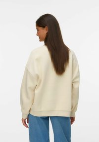 Sweatshirt surdimensionné couleur crème avec épaules tombantes, poignets côtelés et texture douce, associé à un jean bleu ample.