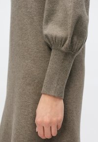 Brązowa dzianinowa sukienka z prążkowanym mankietem i projektowanymi bufkami na rękawach. Miękka tekstura o luźnym kroju, podkreślająca ramię i dłoń.