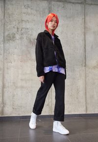 Czarna polarowa kurtka na zamek, fioletowa koszulka z tye-dye pod spodem, czarne spodnie i białe trampki na wysokiej podeszwie. Dodatki to srebrne kolczyki.