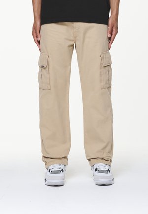 Pantalon cargo beige avec poches à rabat latérales porté avec des baskets athlétiques blanches et noires, debout contre un fond blanc uni.