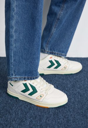 Hvide sneakers med grønne chevron-detaljer, tekstureret læderoverdel, snørelukning og gummisål med orange og teal detaljer; båret med jeans.