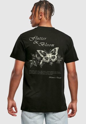 Sort bomulds T-shirt med et stort hvidt sommerfuglegrafik og blomsterdetaljer på ryggen, med teksten "Flutter & Bloom" ovenfor.