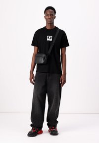 Svart t-skjorte med grafisk trykk, løstsittende mørke jeans og en liten svart crossbody-veske. Røde og svarte joggesko fullfører antrekket.
