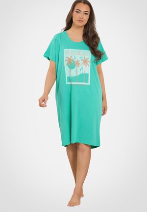 Kobieta w luźnej turkusowej sukience do kolan z krótkimi rękawami i grafiką palmy z przodu, stojąca boso na białym tle.