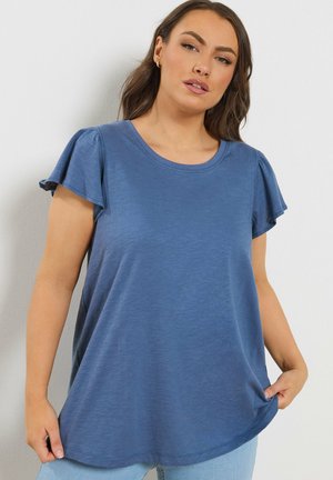 Frau trägt ein locker sitzendes blaues Kurzarmshirt mit flatternden Ärmeln, steht vor einem schlichten weißen Hintergrund.