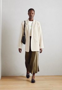 Beige lakan takki valkoisen graafisen t-paidan, oliivivihreän hameen ja mustien loafer-kengien päällä. Mallilla on pieni musta laukku. Taustalla puinen lattia.