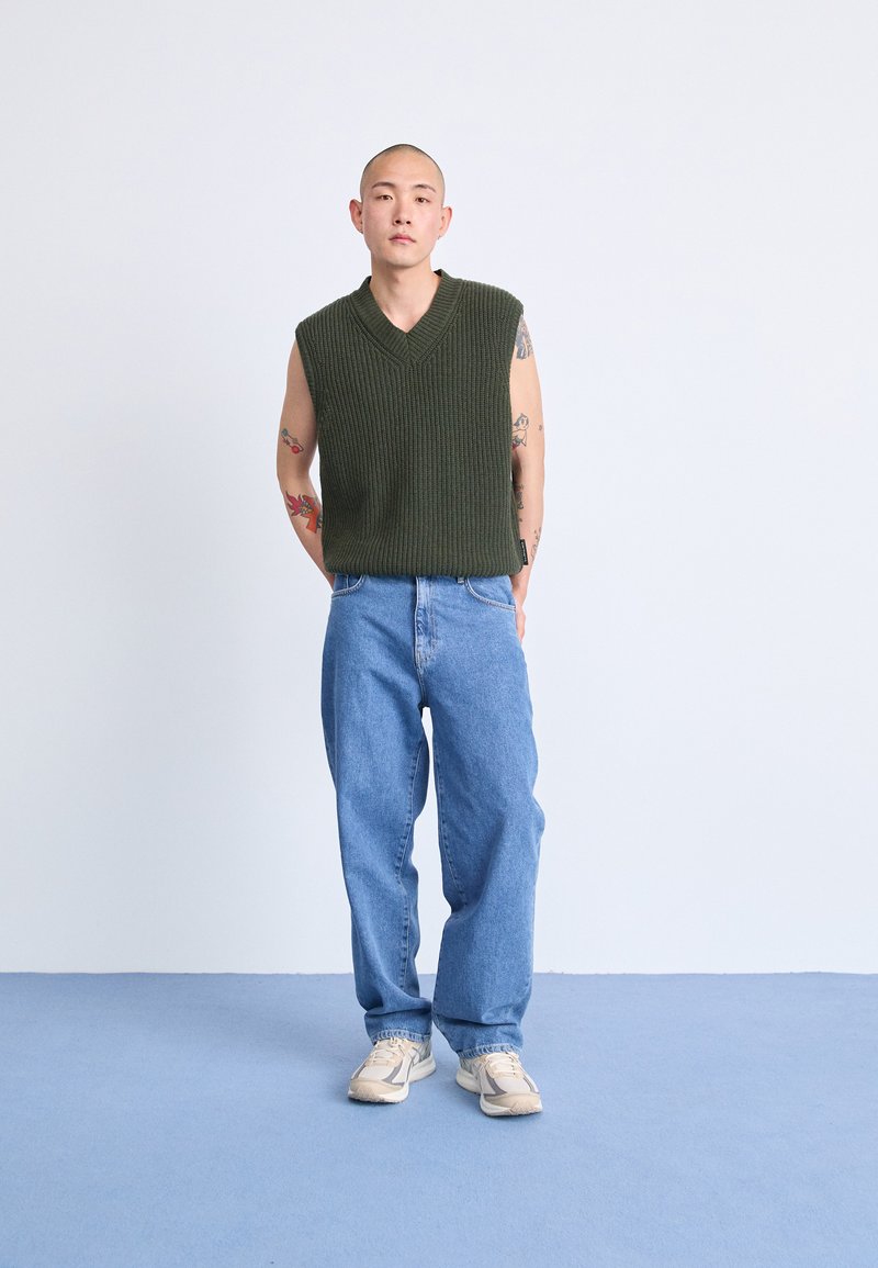 Suéter sin mangas de punto verde con escote en V, combinado con unos jeans de mezclilla de color azul claro. Zapatillas de color claro completan el conjunto, sobre un suelo azul.