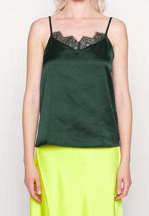 Kobieta ubrana w ciemnozielony satynowy top na ramiączkach z czarną koronkową lamówką oraz neonowożółtą satynową spódnicę, stojąca na jednolitym tle.