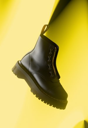 Bottine en cuir noir avec finition texturée, fermeture éclair dorée et bout rond. Présente une semelle épaisse et robuste ainsi qu'une couture jaune contrastante.