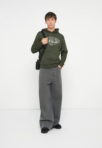 Olivinės spalvos su kapuču palaidinė su balta grafine tekstu, derinama su laisvais pilkais džinsais. Modelis laiko mažą juodą crossbody krepšį ir vilki juodus batus.