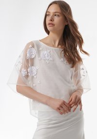 Blusa blanca semitranslúcida con bordado floral; presenta mangas cortas y anchas y un cuello redondo, superpuesta sobre una falda blanca ajustada.