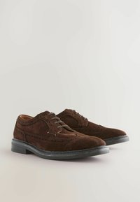 Chaussures oxford en daim marron avec une surface texturée, présentant des détails en forme d'aile et des semelles en caoutchouc noir. Fermeture à lacets avec perforations.