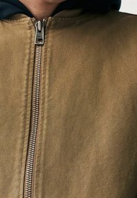 Chaqueta caqui con cremallera, tejido texturizado, cremallera metálica y capucha negra asomando por debajo. Presenta un diseño limpio y costuras resistentes.