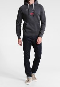 Felpa grigia con logo e design a scudo, tasca a marsupio, coulisse regolabili, indossata con jeans scuri e sneakers beige.