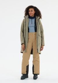Beige parka met een capuchon over een blauwe langetermijn-top, gecombineerd met tan broek met zwarte accenten. Zwarte sneeuwlaarzen. Gladde textuur.