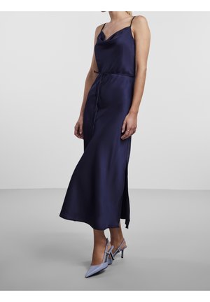 Kobieta w granatowej, satynowej sukience midi bez rękawów, z wciętą talią i bocznym rozcięciem, do tego jasnoniebieskie, ostre szpilki typu slingback.