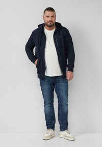 Marine blauwe zip-up hoodie met een voorzak, gelaagd over een wit T-shirt, gecombineerd met blauwe jeans en lichtgekleurde sneakers. Soepel materiaal.