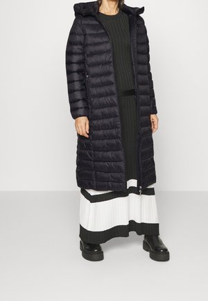 Femme portant un long manteau matelassé noir sur une jupe plissée noire et blanche avec des bottes noires, debout devant un fond uni.
