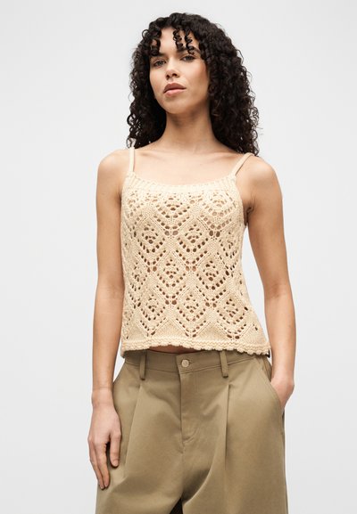 Γυναίκα με κατσαρά μαλλιά που φοράει μπεζ ανάγλυφη αμάνικη μπλούζα και φαρδιά χακι παντελόνια, στέκεται με το ένα χέρι στην τσέπη μπροστά σε λευκό φόντο.