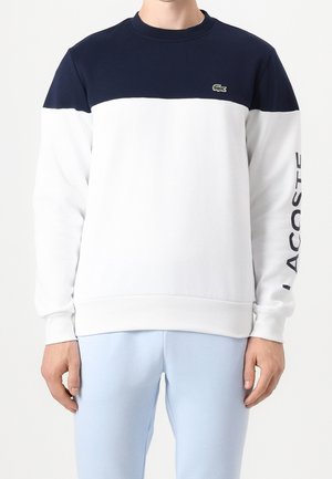 Mann in marineblauem und weißem Sweatshirt mit langen Ärmeln und Lacoste-Logo auf Brust und Ärmel, trägt hellblaue Hose, steht vor weißem Hintergrund.