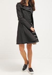 Zwarte dubbelbreasted trenchcoat met een gladde textuur, schouderstukken en zilveren hardware, gelaagd over een zwart-wit gestreepte jurk.