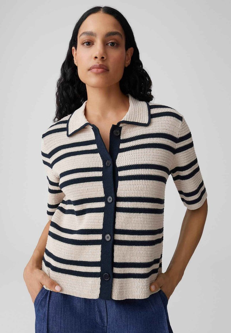 Femme aux longs cheveux foncés portant une chemise à manches courtes beige et marine à rayures boutonnée et un pantalon bleu, les mains dans les poches.