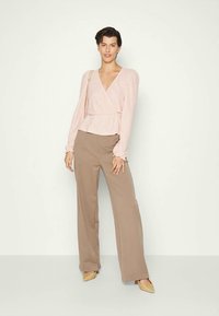 Mulher a usar uma blusa de manga longa rosa claro, calças largas bege, sapatos de salto agudo bege, e a carregar uma bolsa de ombro bege.