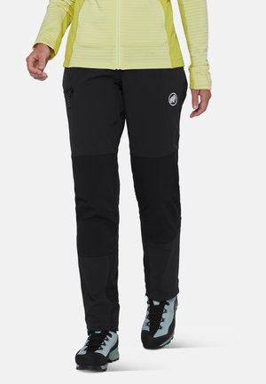 Person trägt schwarze Wanderhose mit Reißverschlusstasche und hellblauen Wanderschuhen, kombiniert mit einer gelben Jacke.