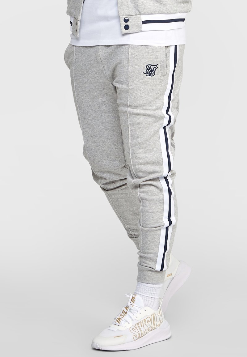 Pantalon de survêtement gris en tissu doux avec une coupe slim, présentant des rayures latérales bleu marine et un logo brodé sur la cuisse. Associé à des baskets blanches.