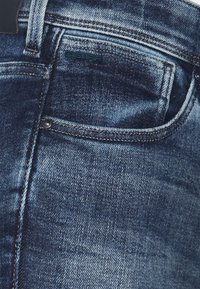 Jeans de denim azul oscuro con un efecto desgastado, que cuentan con un bolsillo trasero, detalles de costura y una pequeña etiqueta de logo cerca de la cinturilla.