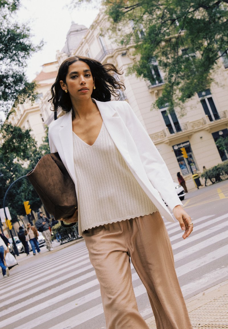 Femme aux longs cheveux foncés traverse la rue en ville, portant un blazer blanc, un haut en maille beige et un pantalon large beige, tenant un grand sac marron.