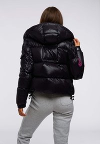 Czarna kurtka puchowa z kapturem, o błyszczącym wykończeniu, pikowanym wzorze i fioletowej naszywce z logo na lewym rękawie. Dopasowany krój.