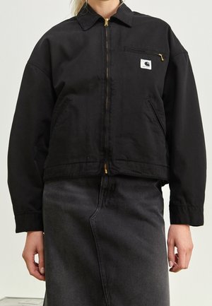 Persona que lleva una chaqueta negra con cremallera, bolsillo en el pecho y logo, combinada con una falda vaquera negra de talle alto.