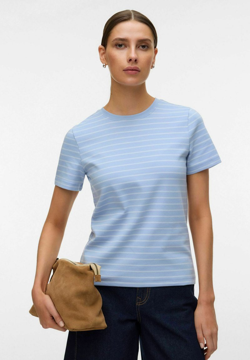 Femme portant un t-shirt à rayures bleu clair et un jean foncé, tenant une pochette en daim marron devant un fond uni.