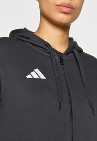 Μαύρο φούτερ με φερμουάρ από λείο ύφασμα, με λευκό λογότυπο με τρεις κάθετες ρίγες στο στήθος και μαύρα κορδόνια στη κουκούλα.