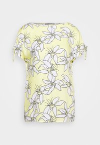 Kortærmet top i lysegult stof med et blomster mønster af hvide blomster med sorte konturer, samt bånd ved ærmeenderne.