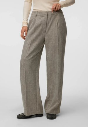 Donna che indossa pantaloni di lana grigia a vita alta con gamba larga e pieghe frontali, abbinati a mocassini neri e top crema a maniche lunghe.