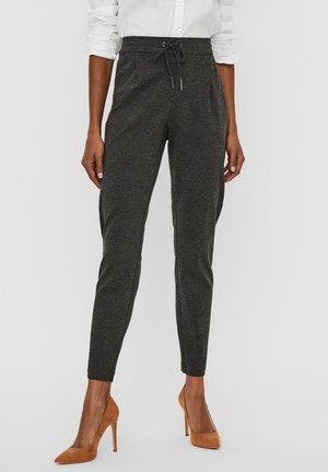 Femme portant un pantalon gris foncé ajusté avec cordon, une chemise blanche aux manches retroussées, et des chaussures à talons marron, debout devant un fond uni.